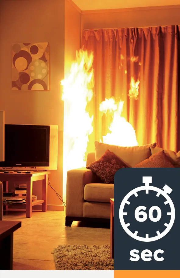 A fire has grown behind a couch in a lounge room and the flames are about reaching almost to the ceiling. The couch and curtains have caught fire.