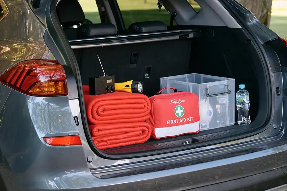 Dark grey car parked on a gravel driveway with the tailgate open. Inside contains emergency supplies including red woolen blankets, a first aid kit, yellow flashlight, portable radio, container with P2 masks and water bottles.